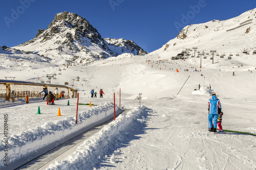Winter resort with children's ski school area with magic carpet conveyor,instructor ans ski slopes between mountains on the background