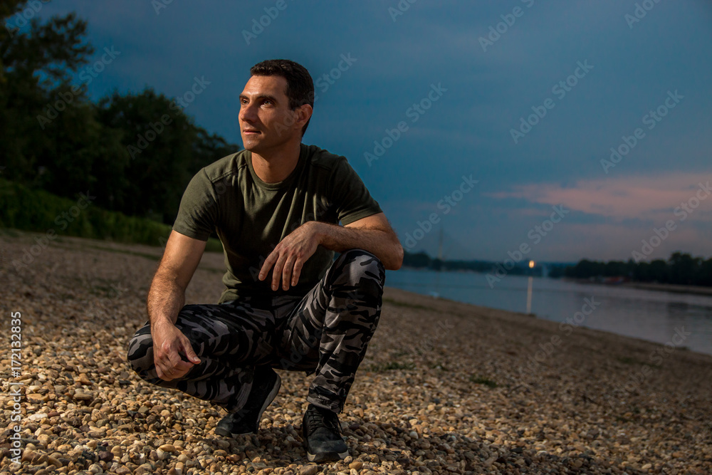 Handsome man in military style seating on rocky beach Handsome man in military style seating on rocky beach