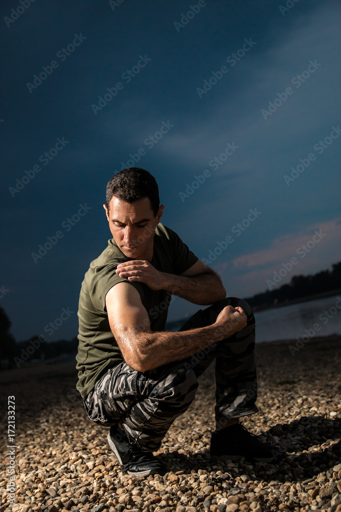 Handsome man in military style demonstrate his muscles and biceps seating on rocky beach