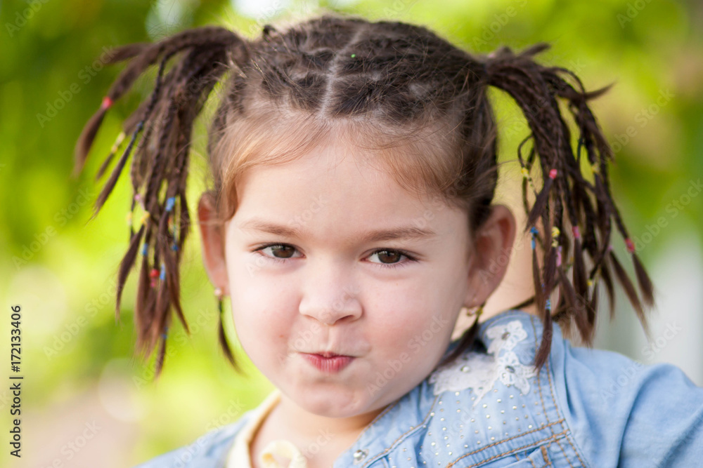 portrait of a little girl with dark eyes, haircut in the form of braids and jeans clothes near a tree on a blurred background portrait of a little girl with dark eyes, haircut in the form of braids and jeans clothes near a tree on a blurred background