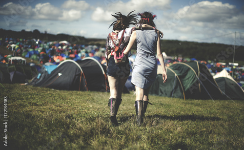 Rear view of two young women at a summer music festival wearing feather headdresses, walking arm in arm towards tents. photo
