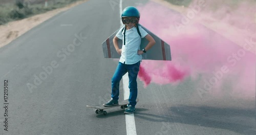 CINEMAGRAPH - seamless loop. Little boy wearing helmet and styrofoam wings standing on a skateboard on a rural road, pretending to be a pilot