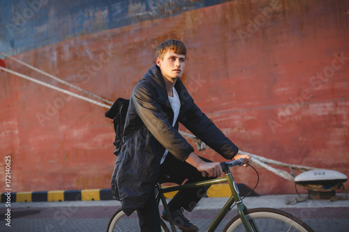 Portrait of young man with brown hair riding classic bicycle while dreamily looking aside. Thoughtful boy in down jacket and black backpack riding bicycle