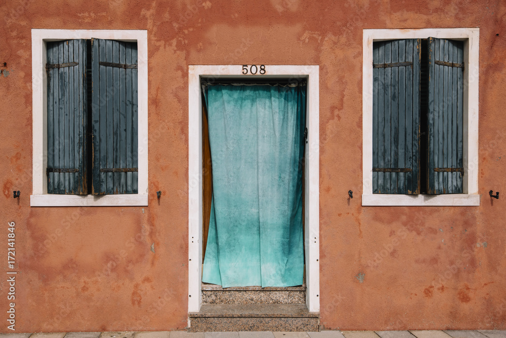 Beautiful colorful house facade on Burano island, north Italy. Red old house wall with a door covered with azure cloth and windows