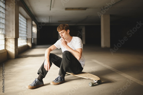 Portrait of boy with brown hair sitting on skateboard and thoughtfully looking down while smoking. Photo of young man in white t-shirt sitting and holding cigarette in hand