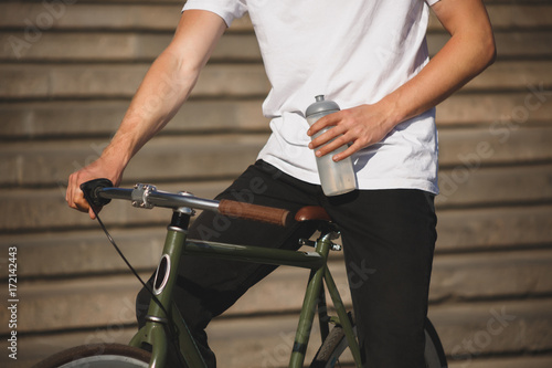 Close up photo of man body in white t-shirt with bicycle and bottle for water in hand. Photo of man body riding a classic bicycle