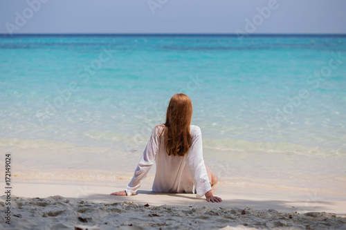 Girl with long red hair sitting with his back on the shore of the blue sea in good weather
