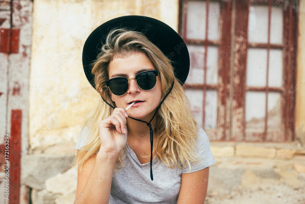 Cocky and confident femme fatale woman with dark vintage sunglasses and cowboy hat, eats or chews lollipop stick in front of wild west style house