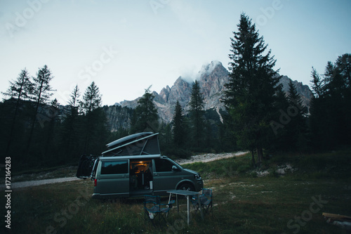 Small travel vehicle camping van or big car with folding rooftop with bed is parked on secluded wild site under huge mountain formation in dolomites, surrounded by forest