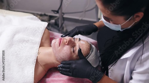 Young woman with blemish skin lying on the couch while professional cosmetologist is applying face cream and making face massage in spa salon