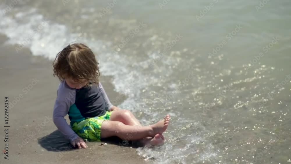 Cute Little Boy Enjoys Playing With A Rock At The Beach, Gentle Waves Crash At His Legs, Slow Motion Cute Little Boy Enjoys Playing With A Rock At The Beach, Gentle Waves Crash At His Legs, Slow Motion