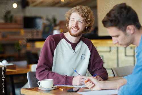 Handsome bearded university student looking away with toothy smile while gathered with his friend at cozy small coffeehouse and preparing for exams, blurred background