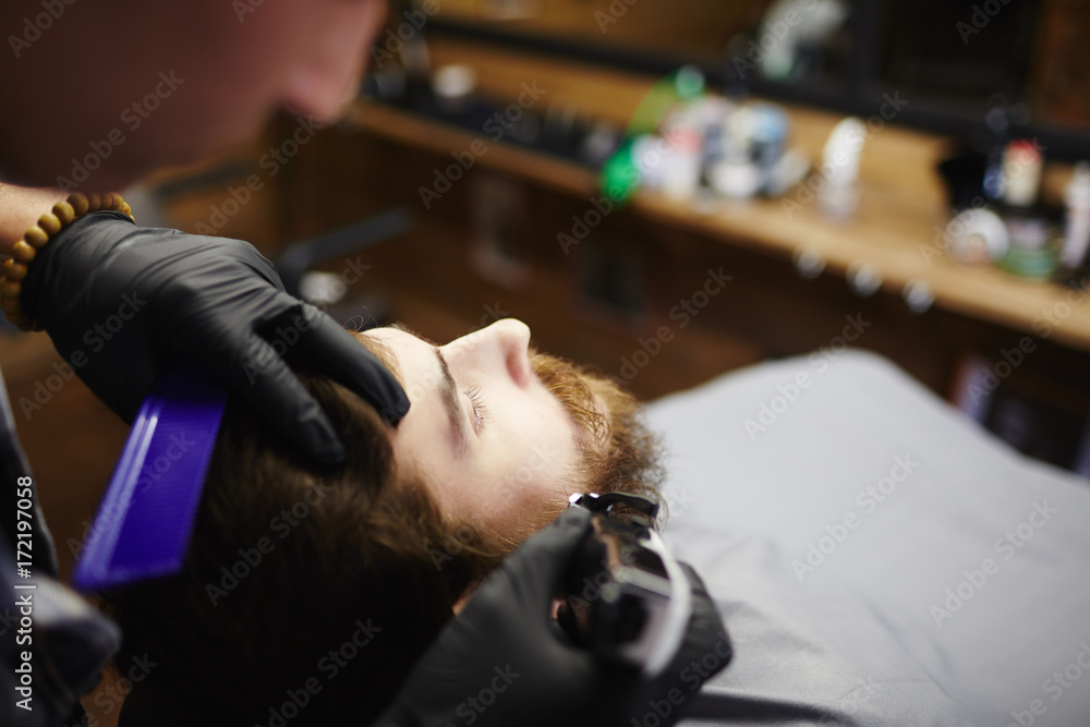 Gloved hands of haircare professional cutting beard of man with electric haircut-machine