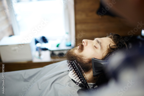 Hairdresser or barber brushing and drying client hair and beard in salon