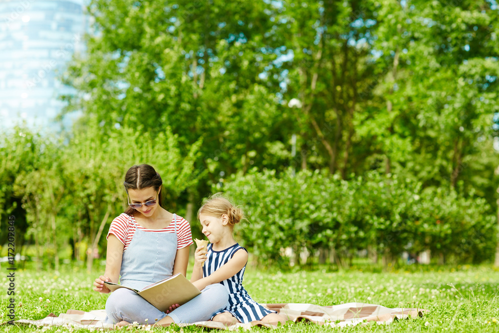 Young woman and her daughter reading book of tales on green lawn on summer day