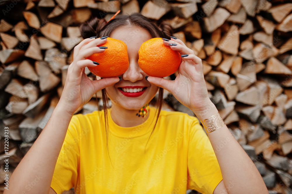 Young funny girl with bright make-up, wear on yellow shirt hold two orange at eyes against wooden background. Young funny girl with bright make-up, wear on yellow shirt hold two orange at eyes against wooden background.