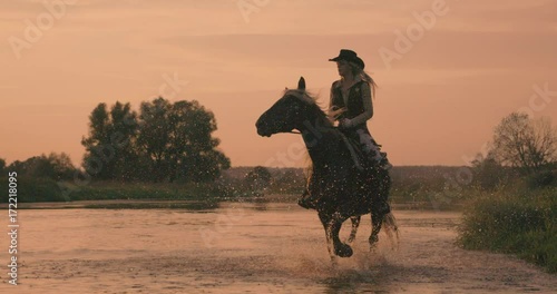 Slow motion 240fps shot of cowboy girl riding horse in water of sunset river