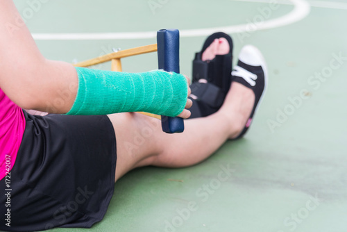 Injured woman wearing sportswear painful arm with gauze bandage, arm cast and wooden crutches sitting on floor.