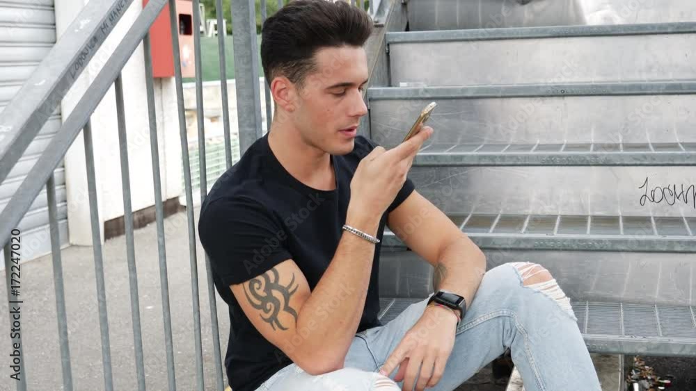 Handsome trendy man wearing white shirt standing and recording voice message on cell phone that he is holding, outdoor in city setting Handsome trendy man wearing white shirt standing and recording voice message on cell phone that he is holding, outdoor in city setting