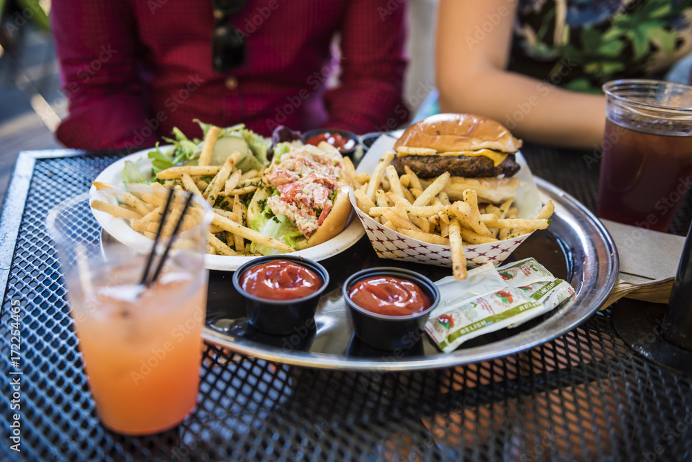 Food, lobster roll, salad, ketchup, relish and burger with fries, and drinks with two people sitting in New York City Food, lobster roll, salad, ketchup, relish and burger with fries, and drinks with two people sitting in New York City