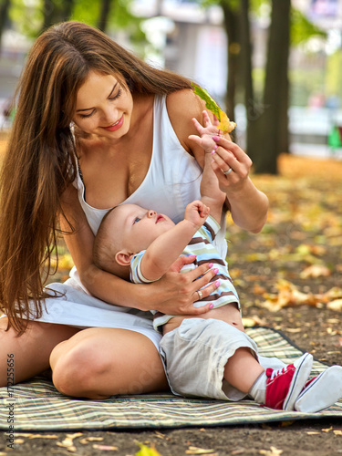 Baby in park outdoor. Kid on mom's hands. Happy beautiful mom and child with leaf sitting on ground in autumn foliage.