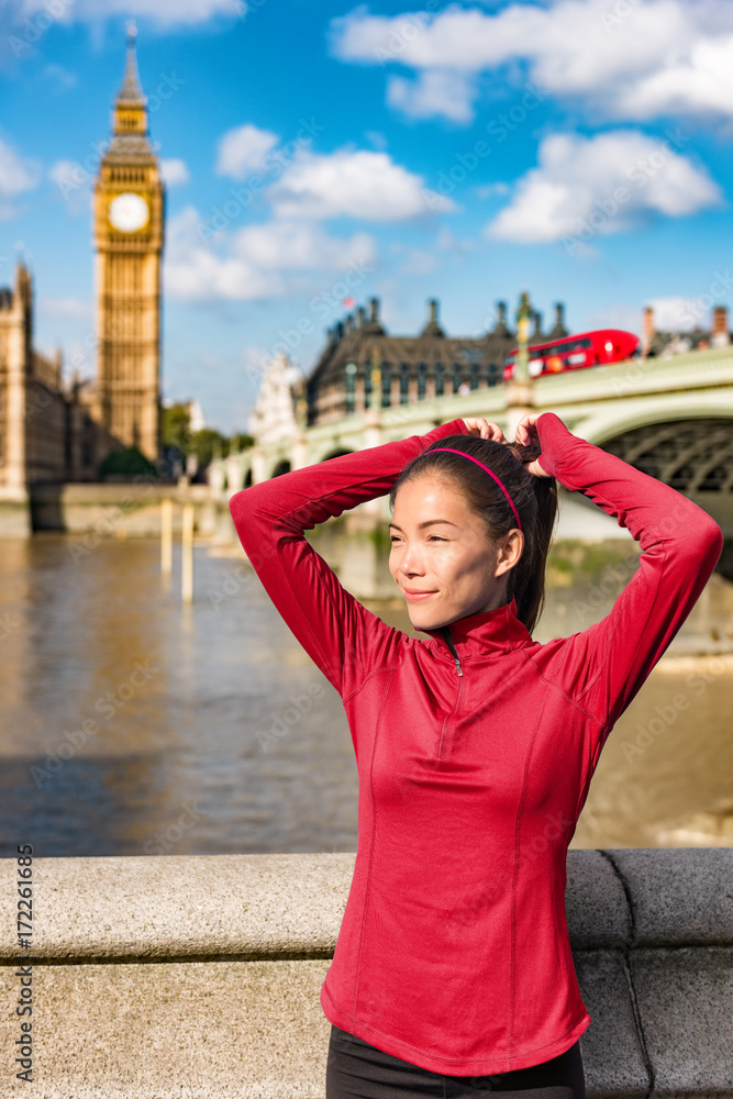 London woman tying hair in ponytail getting ready for autumn run. Beautiful Asian young adult attaching her long brown hair on outdoor running workout at Westminster bridge, London Big Ben, UK.