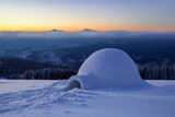 On the snowy lawn in the snowdrift there is an igloo covered by snow with the background of mountains.