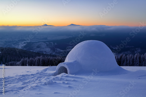 On the snowy lawn in the snowdrift there is an igloo covered by snow with the background of mountains. © Vitalii_Mamchuk