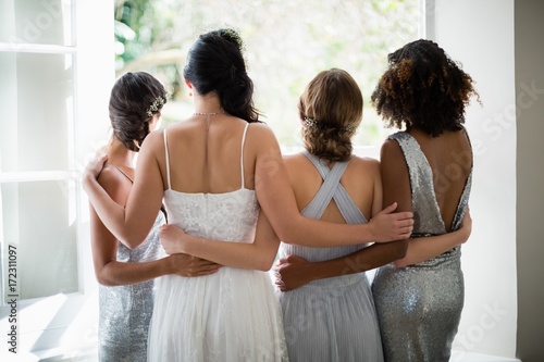 Rear view of bride and bridesmaids standing together near window photo