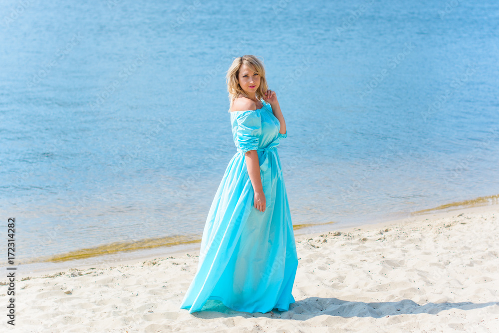 Nice woman in blue long dress walk on a beach and relax at warm day. Tenderness plus size blonde hair lady look stylish and sweet