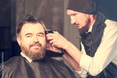 Barber in a beret and a black apron, brushing her hair in front of him sits a bearded man