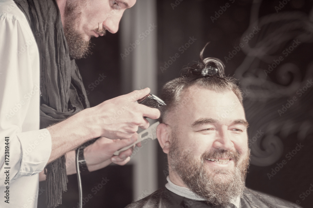 Fototapeta premium Hairdresser in a beret and a black apron cuts with hairclipper a man with a clip on the head