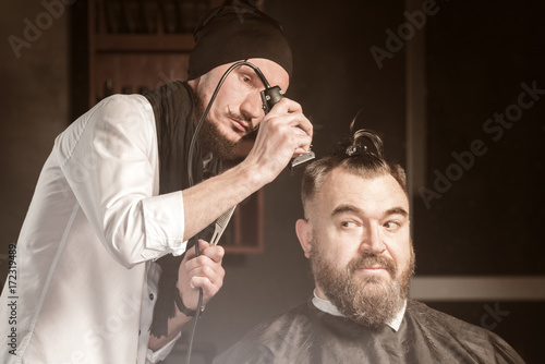 Man hairdresser in a white shirt, black hat and scarf doing hairstyle with hairclipper a bearded man with a clip on the head