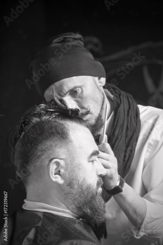 Hairdresser in a black cap and a black apron cuts with hairclipper a man with a clip on the head