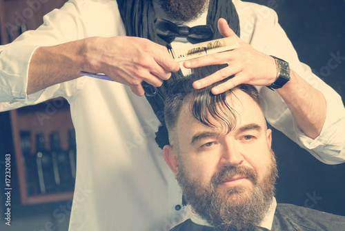 Stylist in black scarf and bow tie makes hair a man with a beard