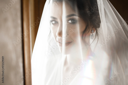 Bride in silk robe and veil stands in the morning lights before a window