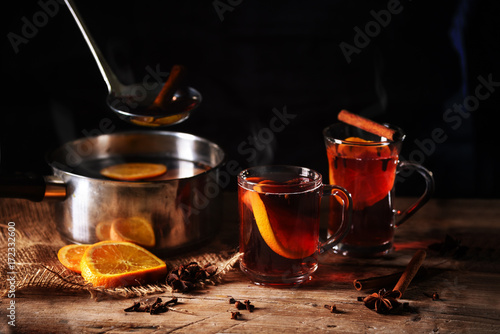 hot steaming mulled wine cooked in a pot and served in glass mugs with Christmas spices like orange slices, cloves, star anise and cinnamon on a rustic wooden table against a dark background