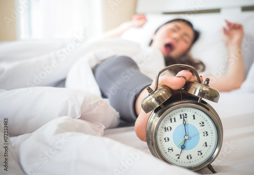 lazy asian beauty woman wake up happy in the morning, foot is stopping alarm clock on white bed in bedroom photo