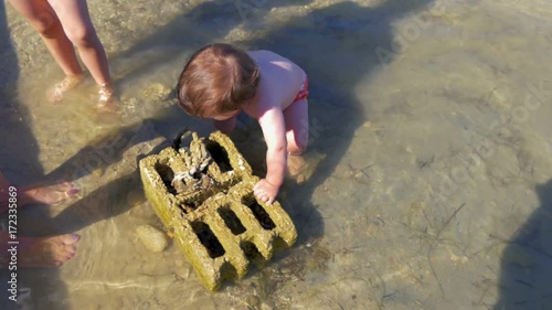 baby boy exploring concreete block laying in shallow sea water. in slow-motion hd photo