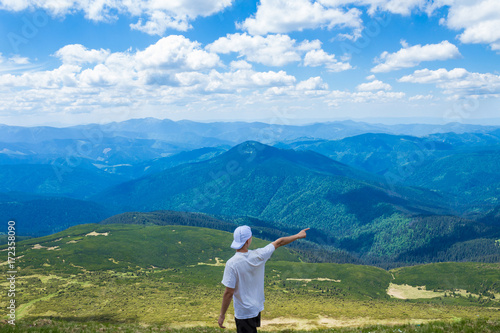 young man in a cap stands at the peak of the mountain with a beautiful view of the mountains and clouds and points to something