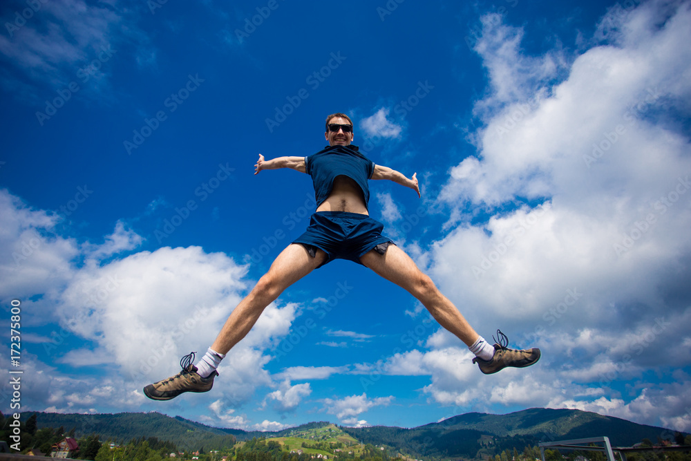 Alone man Jumping for Joy on a Grass Hill above horizon line. Happy slim man fly in a green field against blue summer sky with clouds and mountains. Idea of success, growth, light, diet, luck, freedom Alone man Jumping for Joy on a Grass Hill above horizon line. Happy slim man fly in a green field against blue summer sky with clouds and mountains. Idea of success, growth, light, diet, luck, freedom