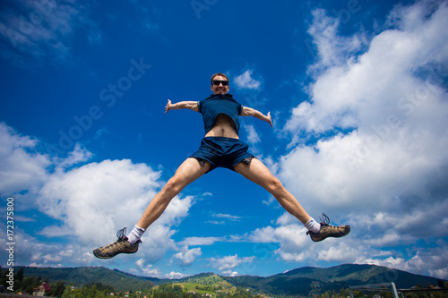 Alone man Jumping for Joy on a Grass Hill above horizon line. Happy slim man fly in a green field against blue summer sky with clouds and mountains. Idea of success, growth, light, diet, luck, freedom © Svetlana