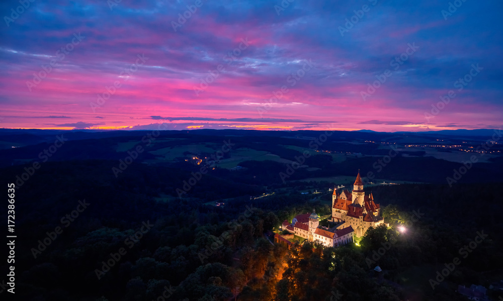 Evening, aerial view on brightly illuminated castle Bouzov on hill against red sunset in picturesque czech landscape. Fairytale, romantic Bouzov castle in late evening, Moravia, Czech republic.
