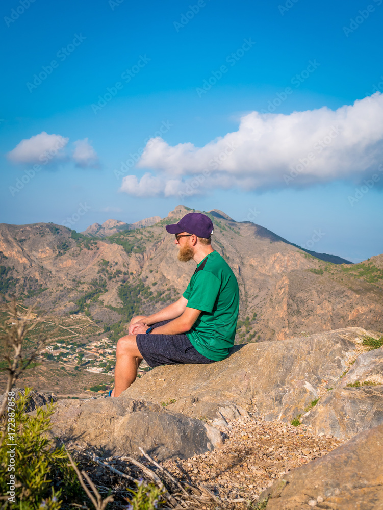 Man with beard and cap sits at the top of a mountain and looking out on the view.