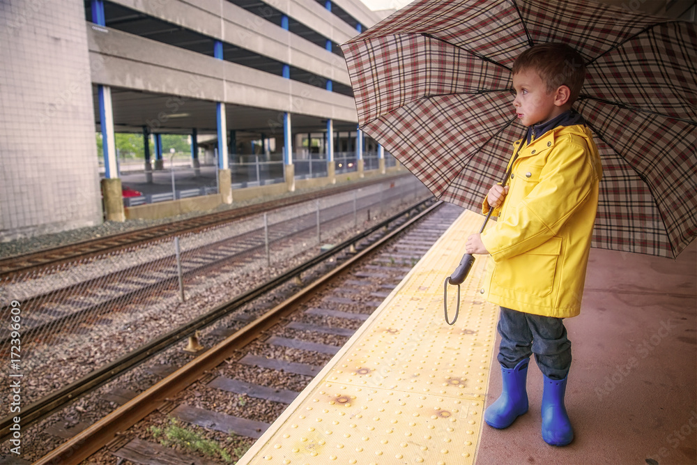 boy at the railway station in rain. cute kid in a yellow raincoat under an umbrella outside. The concept of commute. Copy space for your text