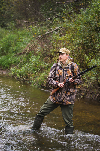 Hunter with a backpack and a hunting gun in the autumn forest. The man is on the hunt. The hunter crosses the forest river.