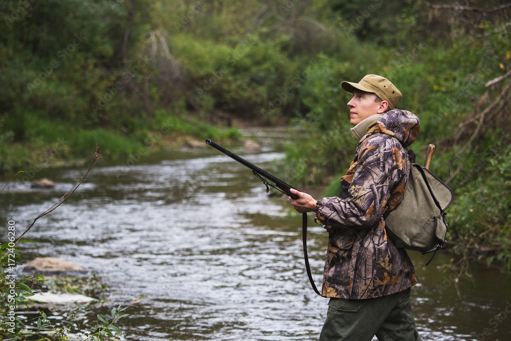 Hunter with a backpack and a hunting gun in the autumn forest. The man is on the hunt. The hunter crosses the forest river. Hunter with a backpack and a hunting gun in the autumn forest. The man is on the hunt. The hunter crosses the forest river.