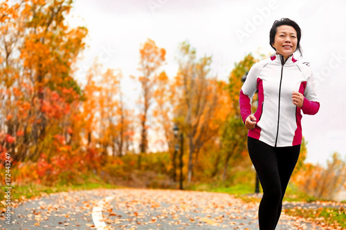Mature happy middle age woman jogging outside in her 50s. Middle aged Asian chinese girl in her fifties jogging outdoor living healthy lifestyle in beautiful autumn city park in colorful fall foliage. photo
