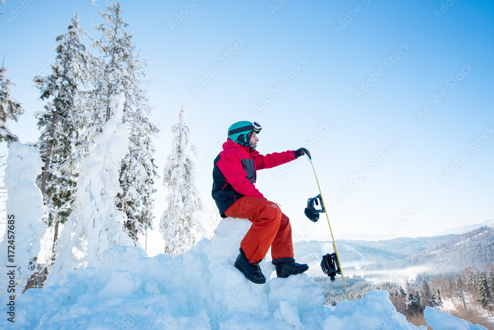 Man snowboarder sitting on top of a snowy hill with his snowboard enjoying stunning mountains view ski resort copyspace seasonal activity. Ski season and winter sports concept