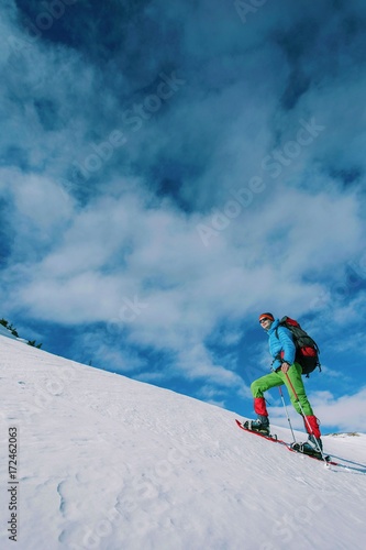 Climber with backpack trekking poles snowshoes rises to the top of the mountain in the snow on a background of beautiful blue sky. 